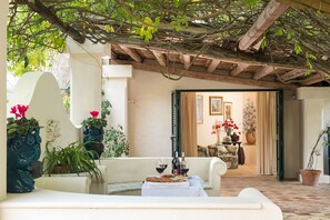 An outdoor seating area with a table set for two, a potted plant, and a view into a room with a couch and a painting. - Belvilla by OYO San Ciro Otto (Salemi)
