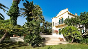 Une maison à deux étages, à l’extérieur blanc et aux volets verts, entourée de palmiers luxuriants et de plantes en pot.