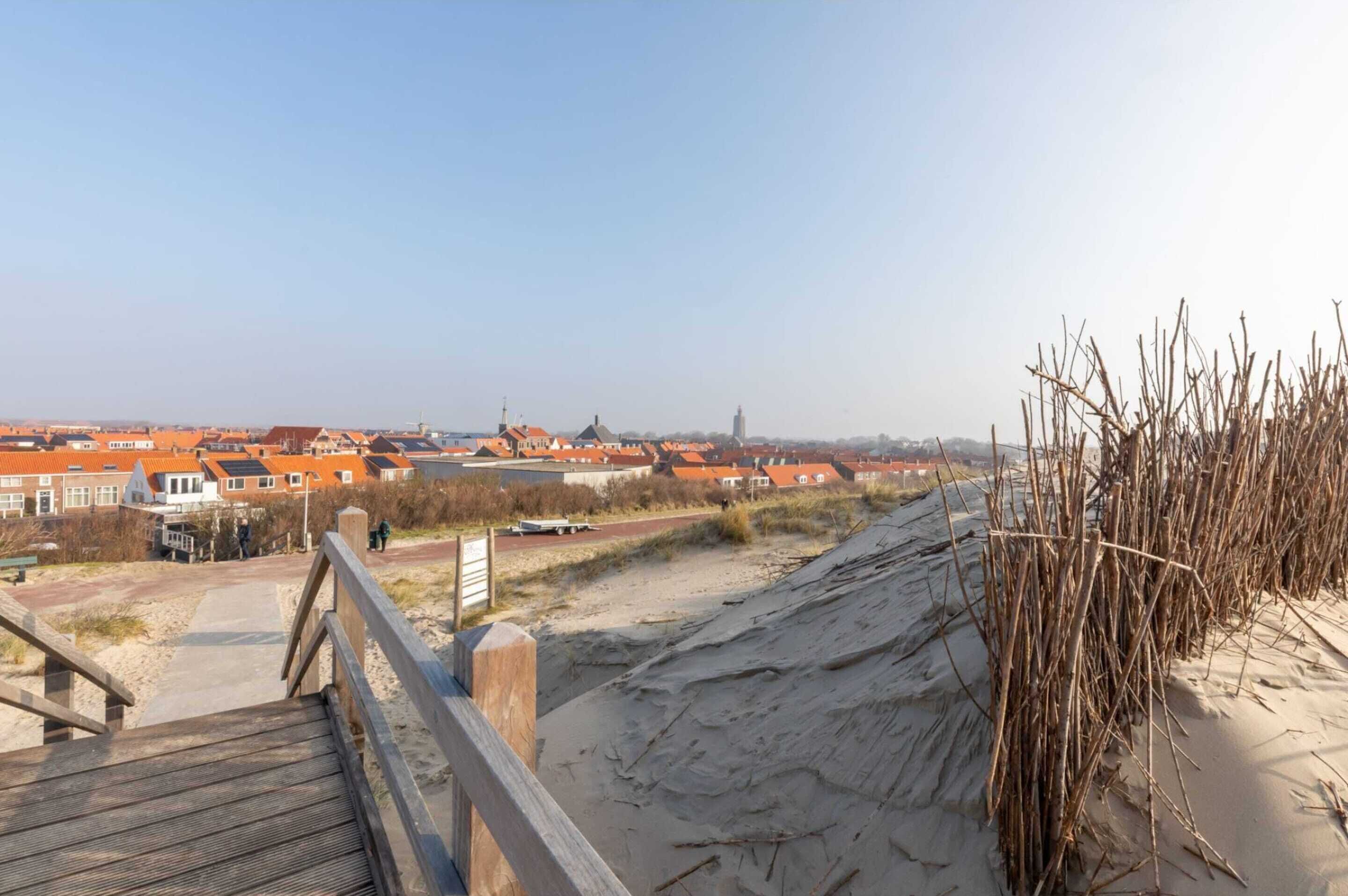 A wooden boardwalk leading to a sandy beach area with a view of a town with orange-roofed buildings.