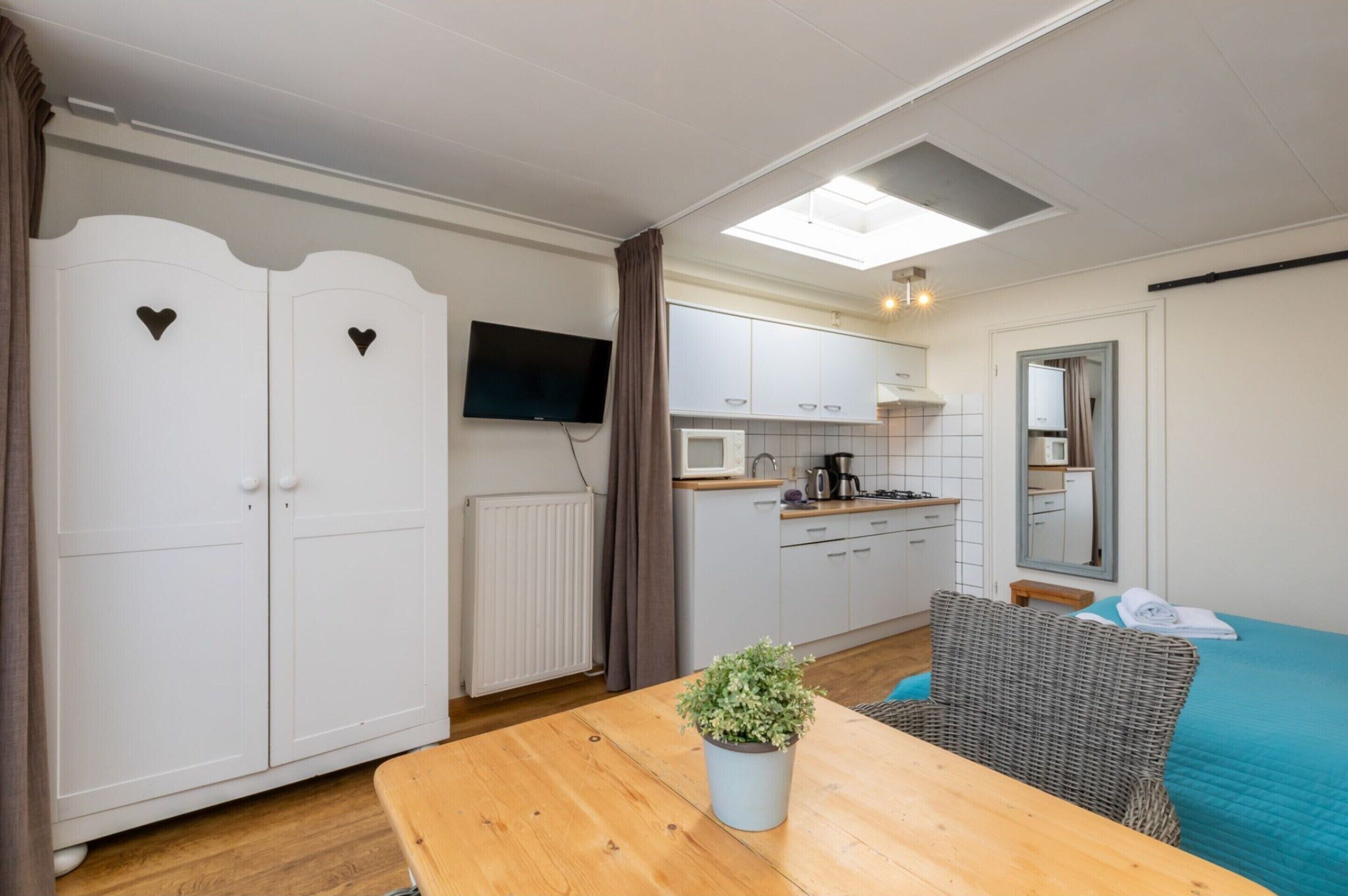 Une pièce compacte avec une table en bois, une plante en pot, une chaise en osier, un lit recouvert d’une courtepointe bleue, un coin cuisine équipé d’un four à micro-ondes, une télévision et une armoire ornée de découpes en forme de cœur.