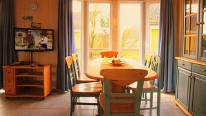 Un comedor con una mesa y sillas de madera, un televisor sobre un mueble de madera, y vistas a través de amplias ventanas.