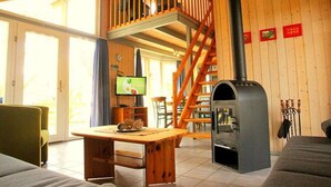 Interior acogedor de una cabaña con una mesa de comedor de madera, un sofá, un televisor, una chimenea y una escalera de madera.