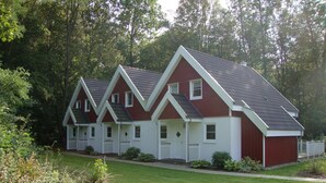 A red and white two-story house with a sloped roof, surrounded by a well-maintained lawn and dense trees. - XXL Ferienhaus in Bad Saarow (Bad Saarow)