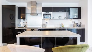 Une cuisine moderne avec des meubles noirs, une hotte en acier inoxydable, un four encastré et un plan de travail sur lequel se trouve un grille-pain. Deux chaises sont placées devant le plan de travail.
