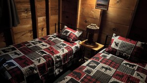 Une pièce chaleureuse avec deux lits, chacun recouvert d’une courtepointe rouge et noire ornée de motifs animaliers. On y trouve un mur en bois et une petite table avec une lampe.