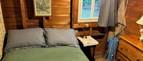 Habitación de cabaña de madera con una cama, una mesita con una lámpara, un tocador y una ventana con vistas a la vegetación.