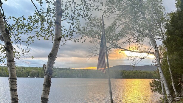 Un feu de camp au bord du lac, avec un drapeau, entouré de bouleaux et d’une jetée en bois.