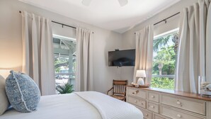 Une chambre à coucher avec un lit, une commode, une chaise, un téléviseur et une fenêtre avec des rideaux.