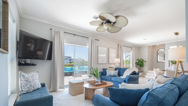 Un salon moderne doté d’un grand téléviseur, de canapés bleus, d’un ventilateur de plafond et offrant une vue sur une piscine.