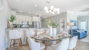 Une cuisine moderne avec des meubles blancs, un réfrigérateur en acier inoxydable et un coin repas comprenant une table en verre et des chaises blanches.