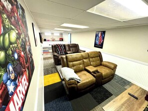 Ein Zimmer mit einer braunen Ledersofa, einem Gaming-Sessel, einem großen Marvel-Poster und einem kleinen Tisch mit einer Fernbedienung.
