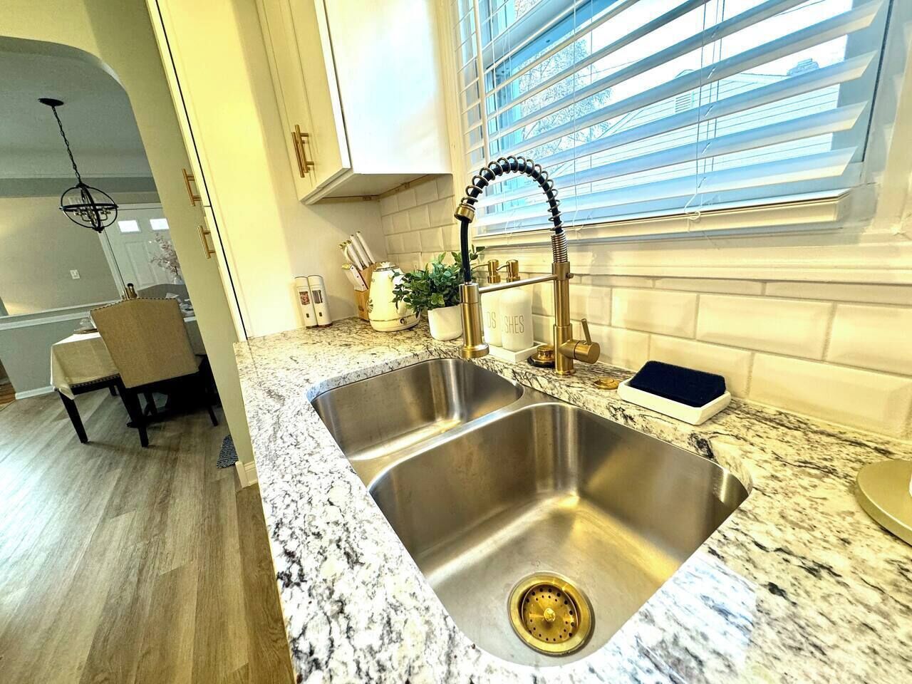 A kitchen with a double stainless steel sink, granite countertop, and a window with blinds.