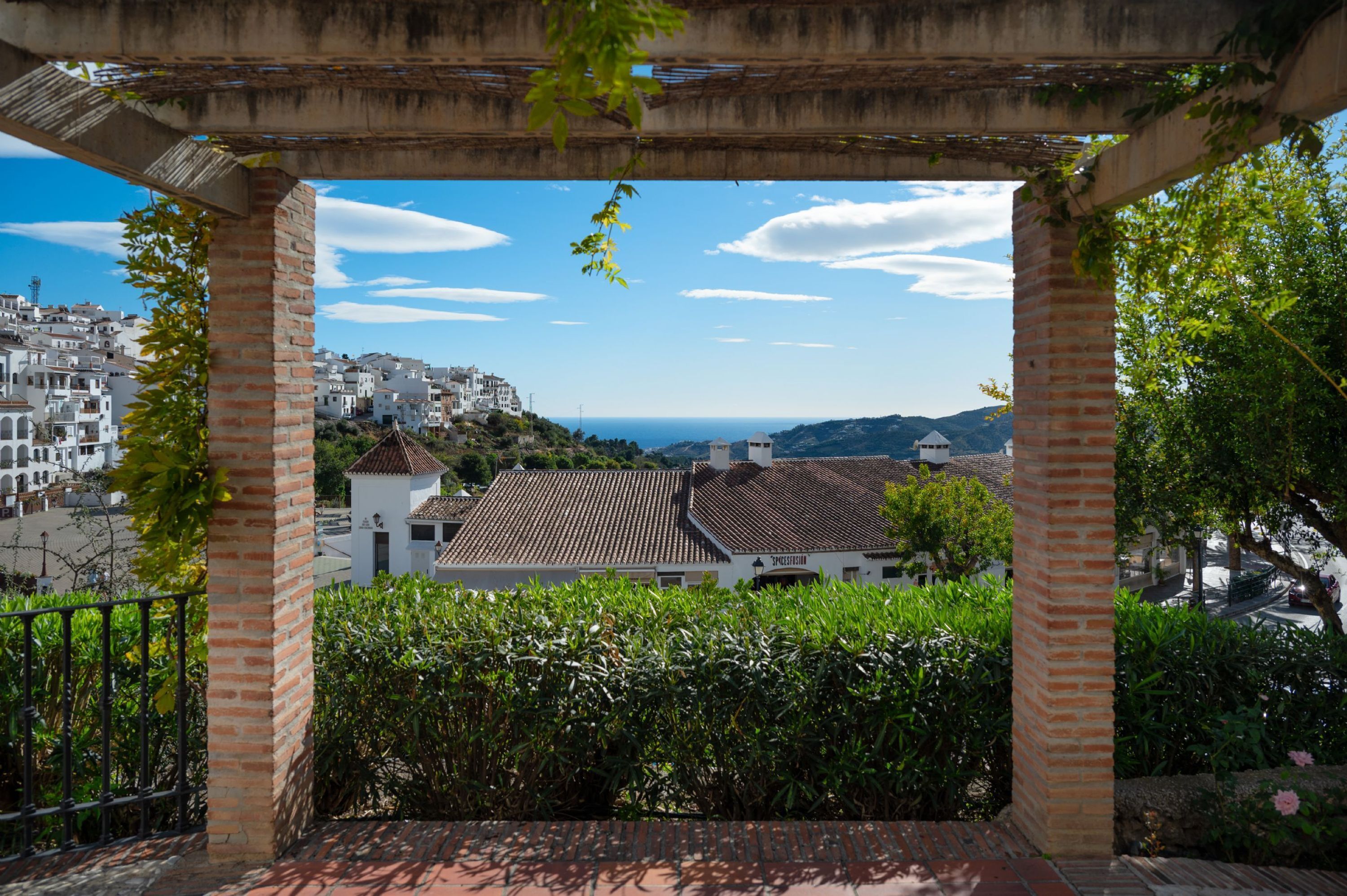 A view from a covered outdoor area with a brick column structure, overlooking a coastal town with white buildings and a clear sky.