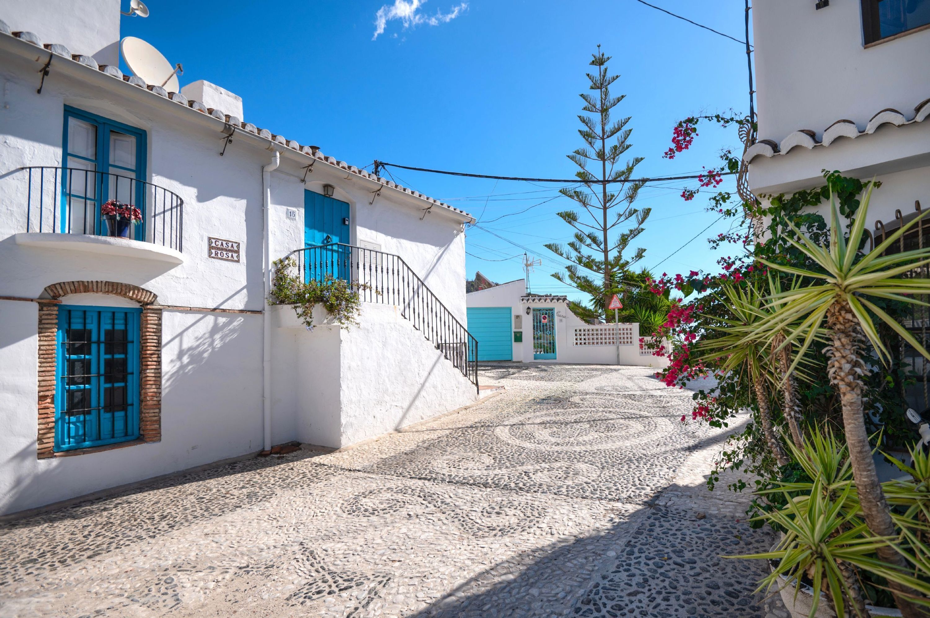 Uma rua de paralelepípedos estreita, ladeada por edifícios brancos com portas e janelas azuis.