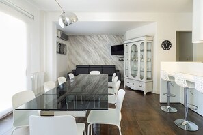 Ett modernt matrum med ett glasbord och vita stolar, en vägg i marmor, en tv och ett skåp med glasdörrar.