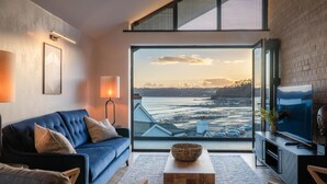 A modern living room with a large window, a blue sofa, a wooden coffee table, and a TV on a black stand. - Wisemans House Apartment 3 - Wisemans Bridge (Saundersfoot)