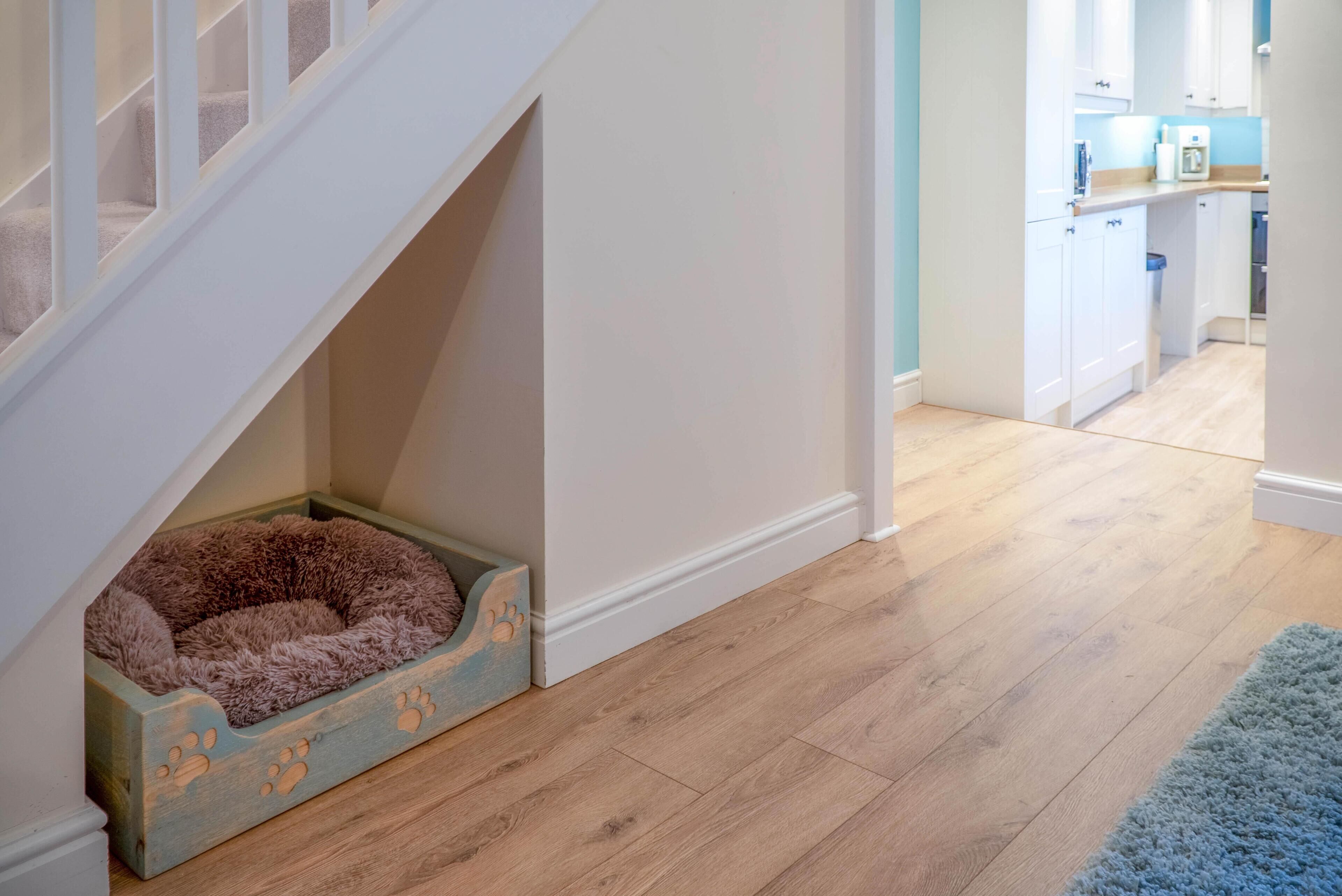 Una escalera con una cama para mascotas al pie, una zona de cocina con armarios y encimera, y suelo de madera.