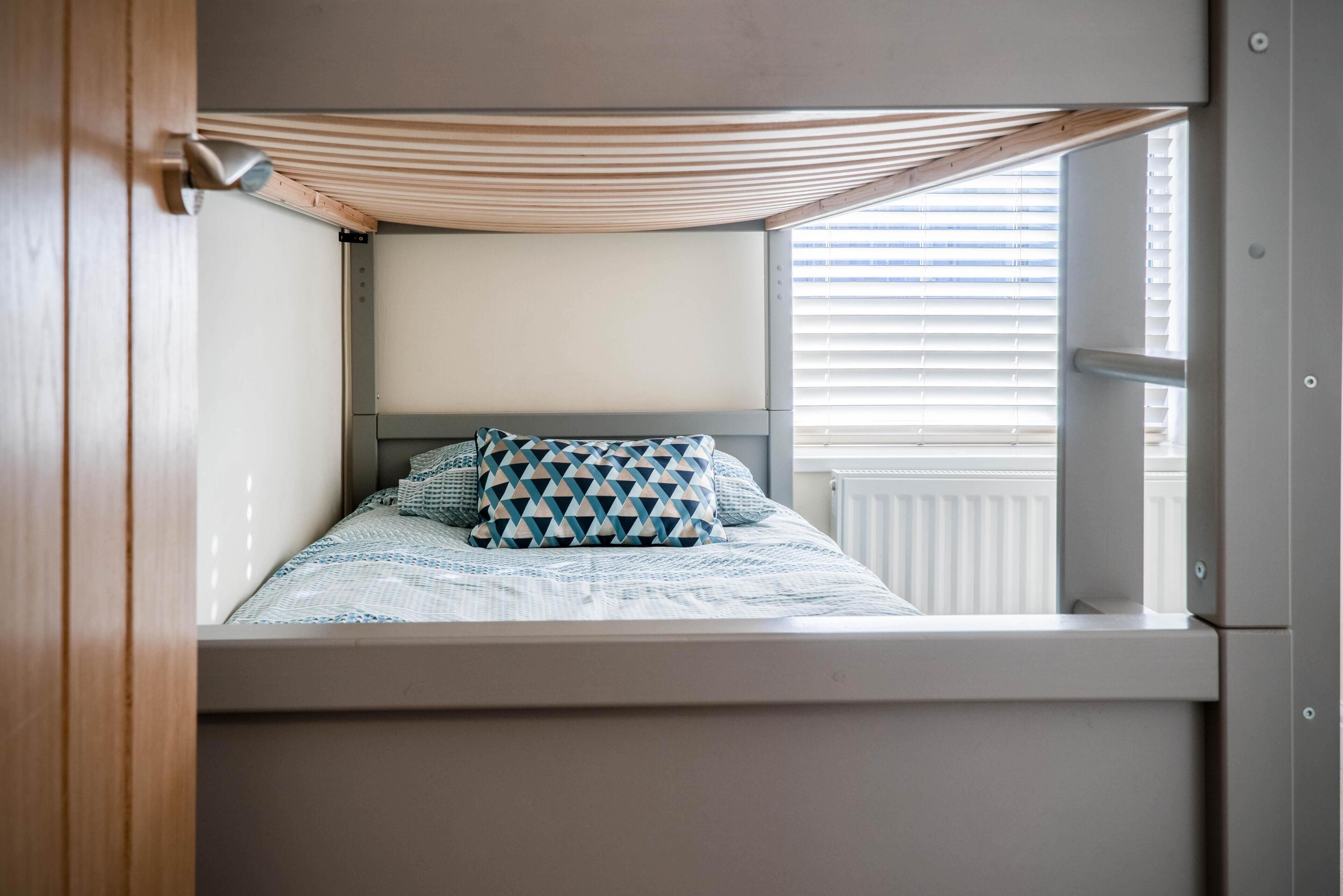 Un lit superposé avec un oreiller à motifs géométriques, une fenêtre avec des stores et un plafond en lattes de bois.