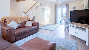 A living room with a brown leather sofa, ottoman, and a television on a wooden stand. - Ty George - 3 Bedroom Cottage - Tenby (Tenby)