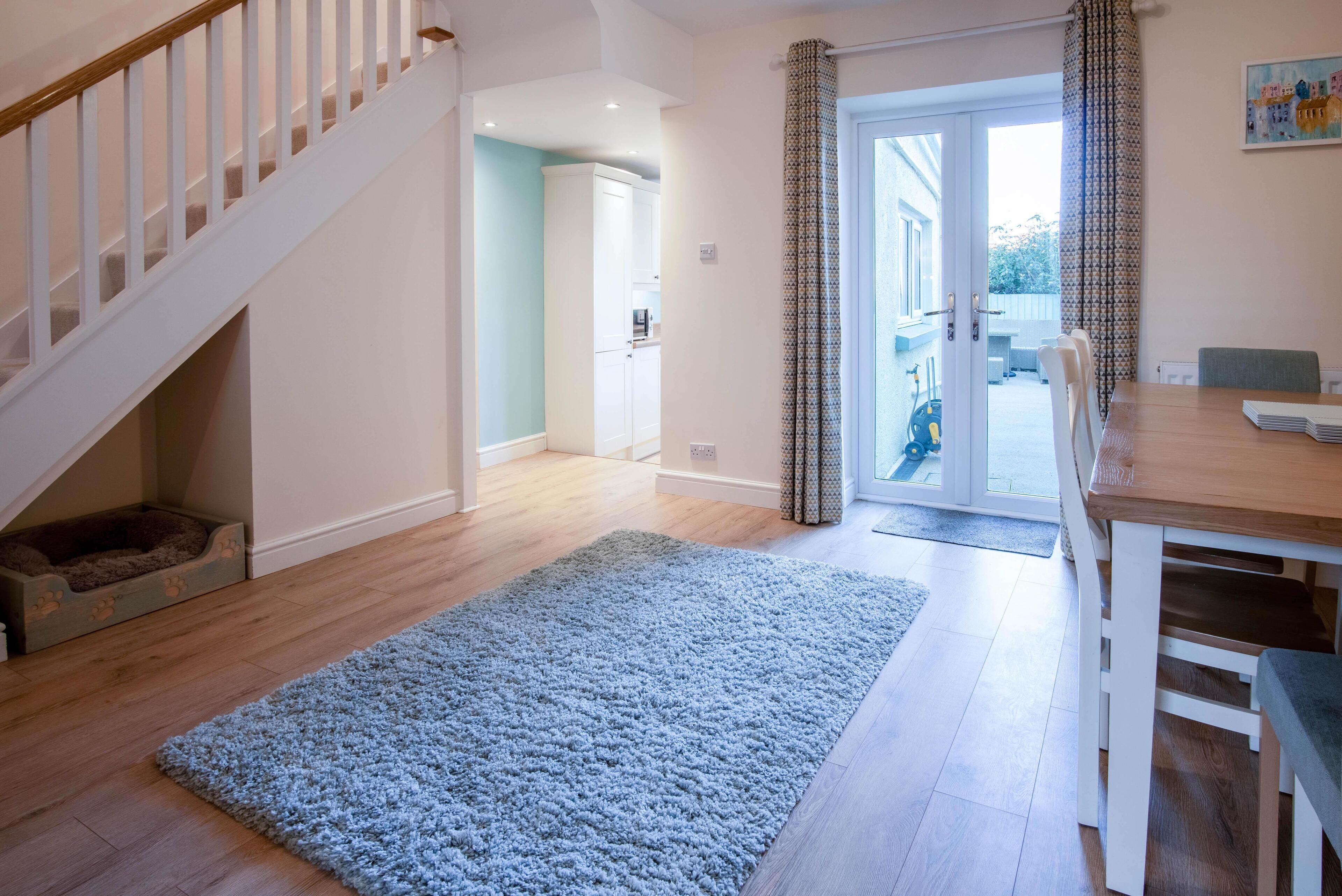 A spacious room with a wooden floor, a blue rug, a dining table with chairs, a staircase, and a door leading outside.