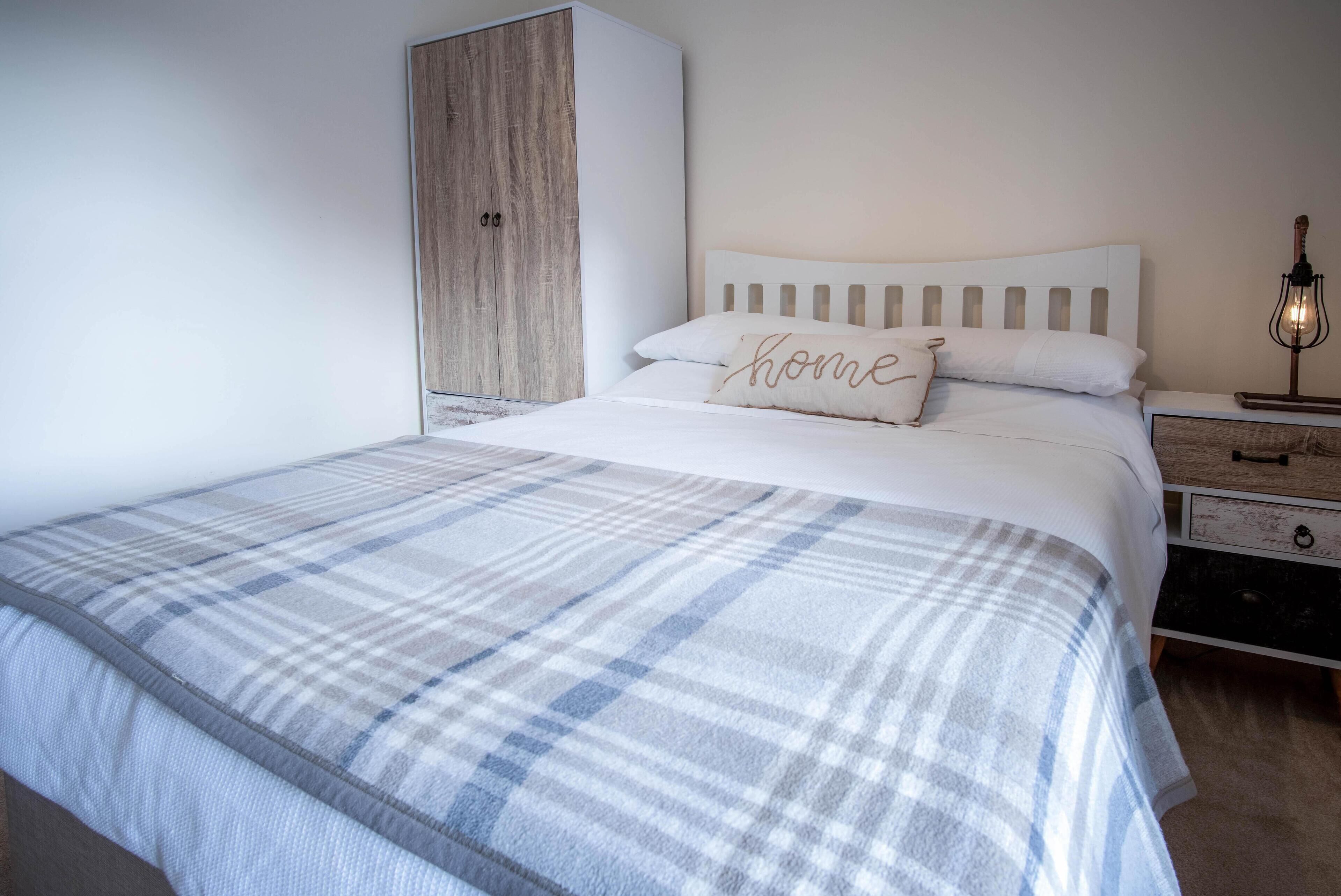 Un lit bien fait, recouvert d’une couverture à carreaux, un oreiller orné du mot « chez soi », une armoire en bois et une lampe de chevet.