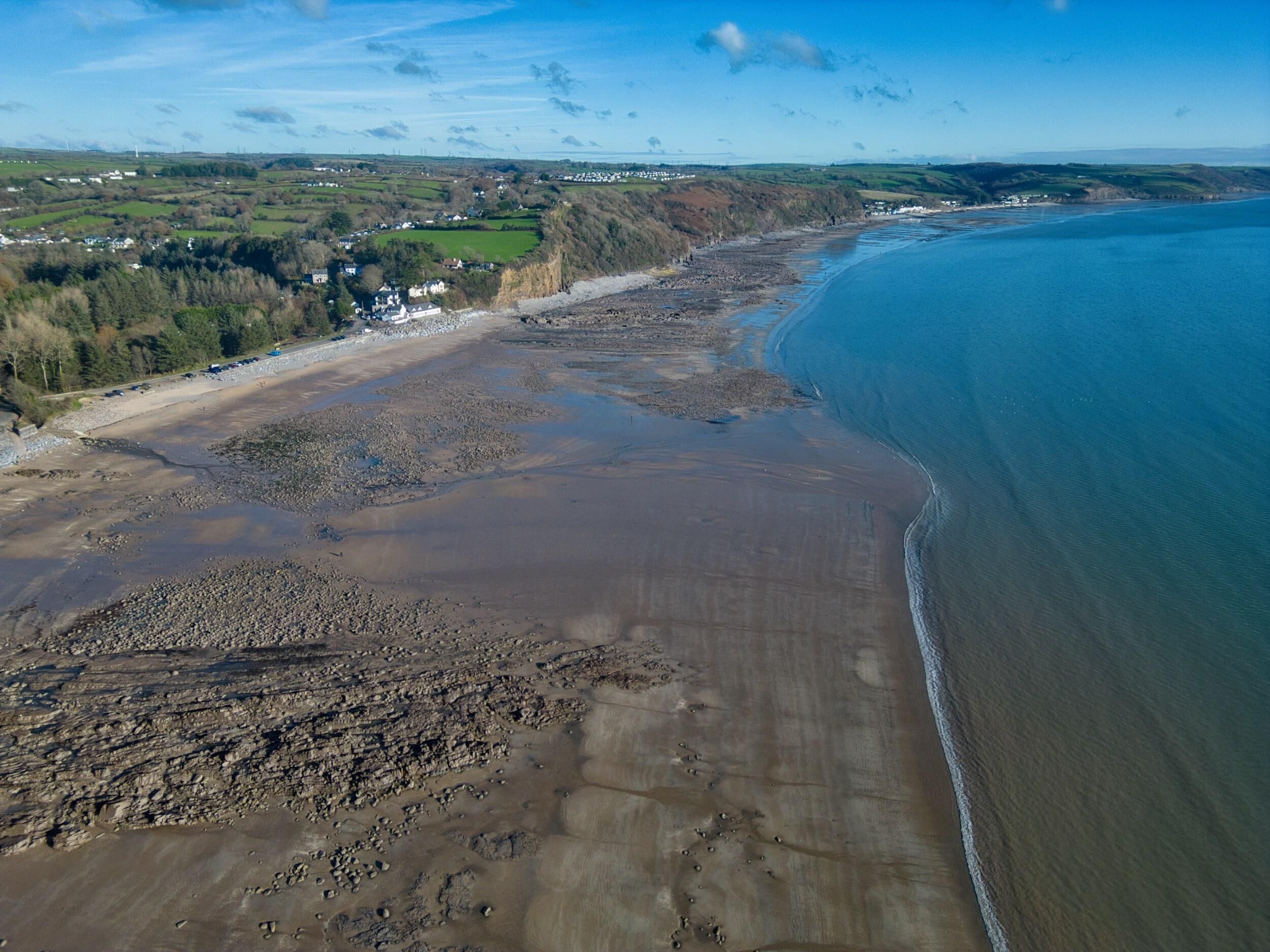 Eine Luftaufnahme einer Küstenlandschaft mit Strand, felsigem Ufer und einem Dorf in der Ferne.