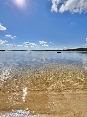 A clear beach with calm water, a distant shoreline, and a partly cloudy sky.