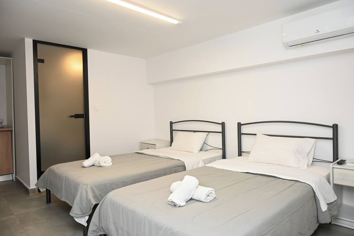 A clean, modern hotel room with two single beds, a bedside table with a lamp, and a built-in air conditioning unit.