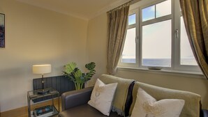 Een woonkamer met een bank, een bijzettafeltje met een lamp, een potplant en uitzicht op de zee door een raam.