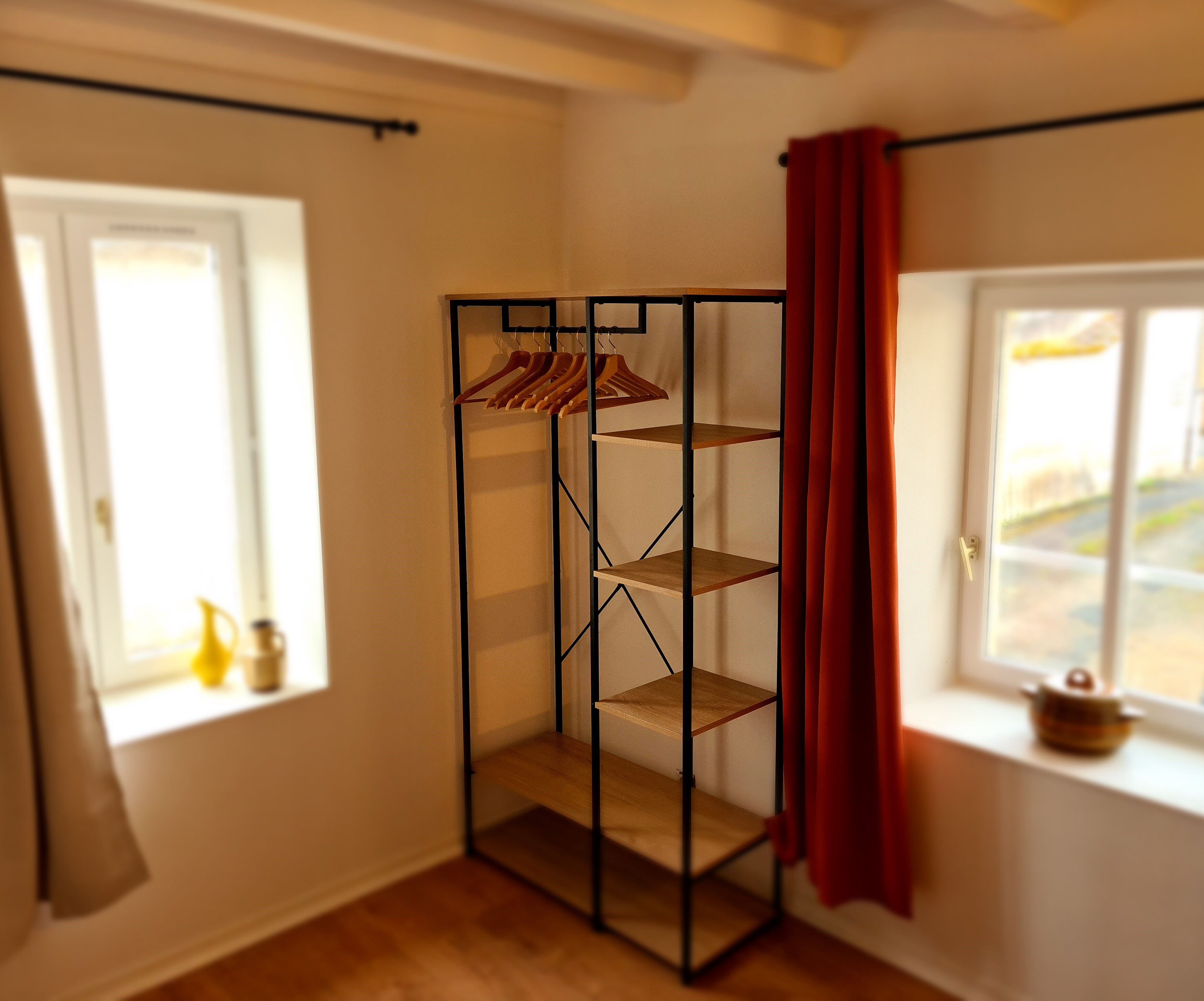 Une pièce avec un plancher en bois, une étagère en métal noir avec des cintres en bois, et un rideau rouge.