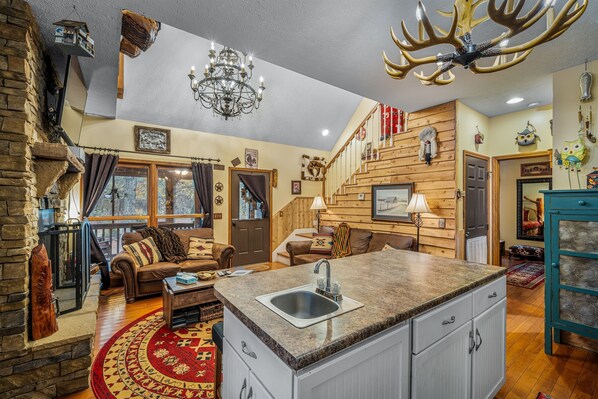 A cozy living room with a fireplace, a large window, and a kitchen area with a granite countertop and sink. - Cedar Cove-fishing, hot tub, fire pit & playhouse (Mentone)