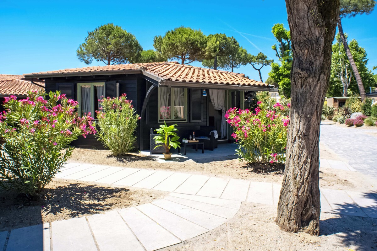 A small house with a tiled roof, a patio with a table and chairs, surrounded by flowering plants and trees.