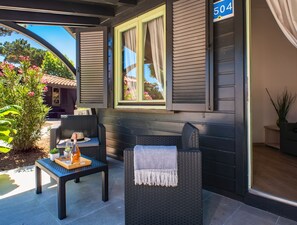 A covered outdoor seating area with wicker furniture, a table with a drink and a hat, and a window with curtains. - Hotiday Milano Marittima Pineta (Cervia)