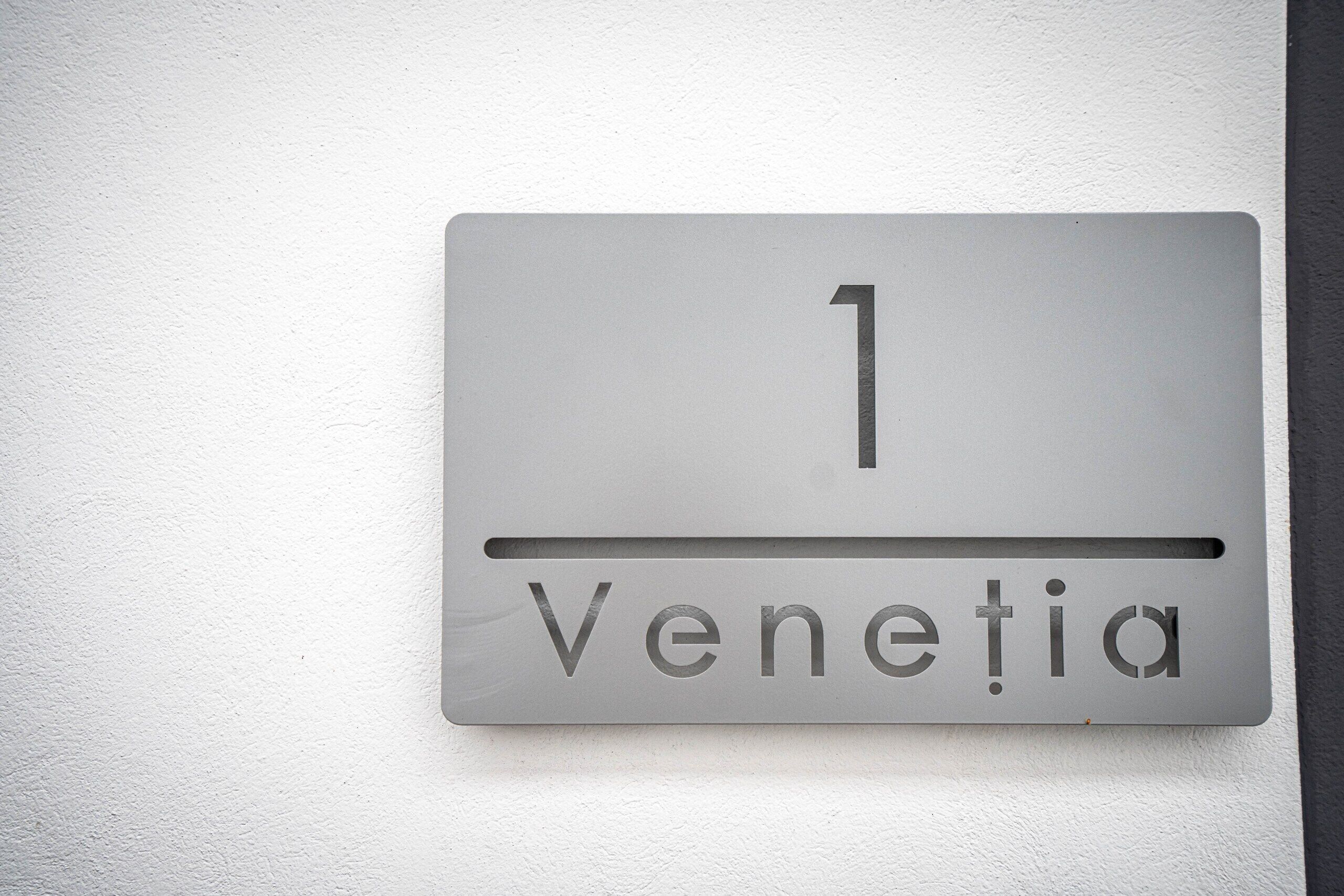 Ein Metallschild mit der Nummer 1 und dem Wort 'Venetia' an einer weißen Wand.