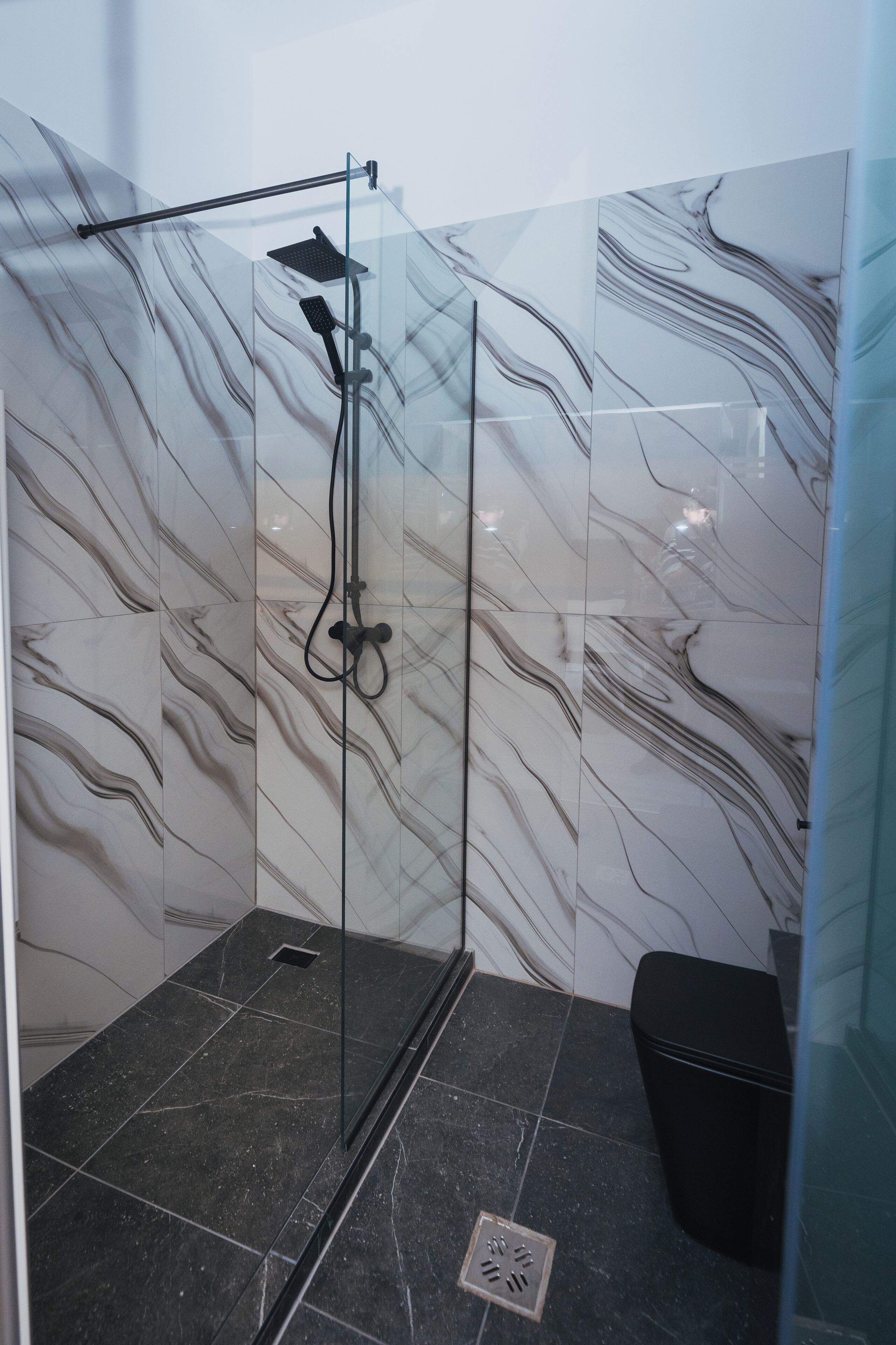 A glass-enclosed shower with a handheld showerhead, a black trash bin, and marble-patterned walls.