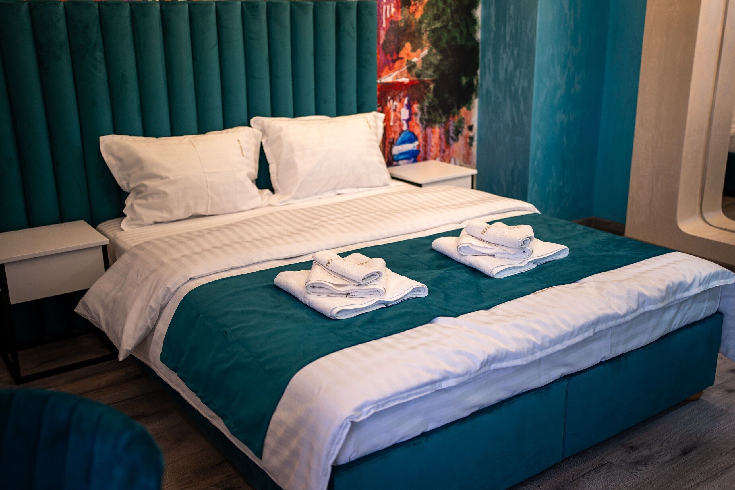 A neatly made bed with white linens and a teal bedspread, a pair of folded towels on the bed, and a white nightstand beside it.