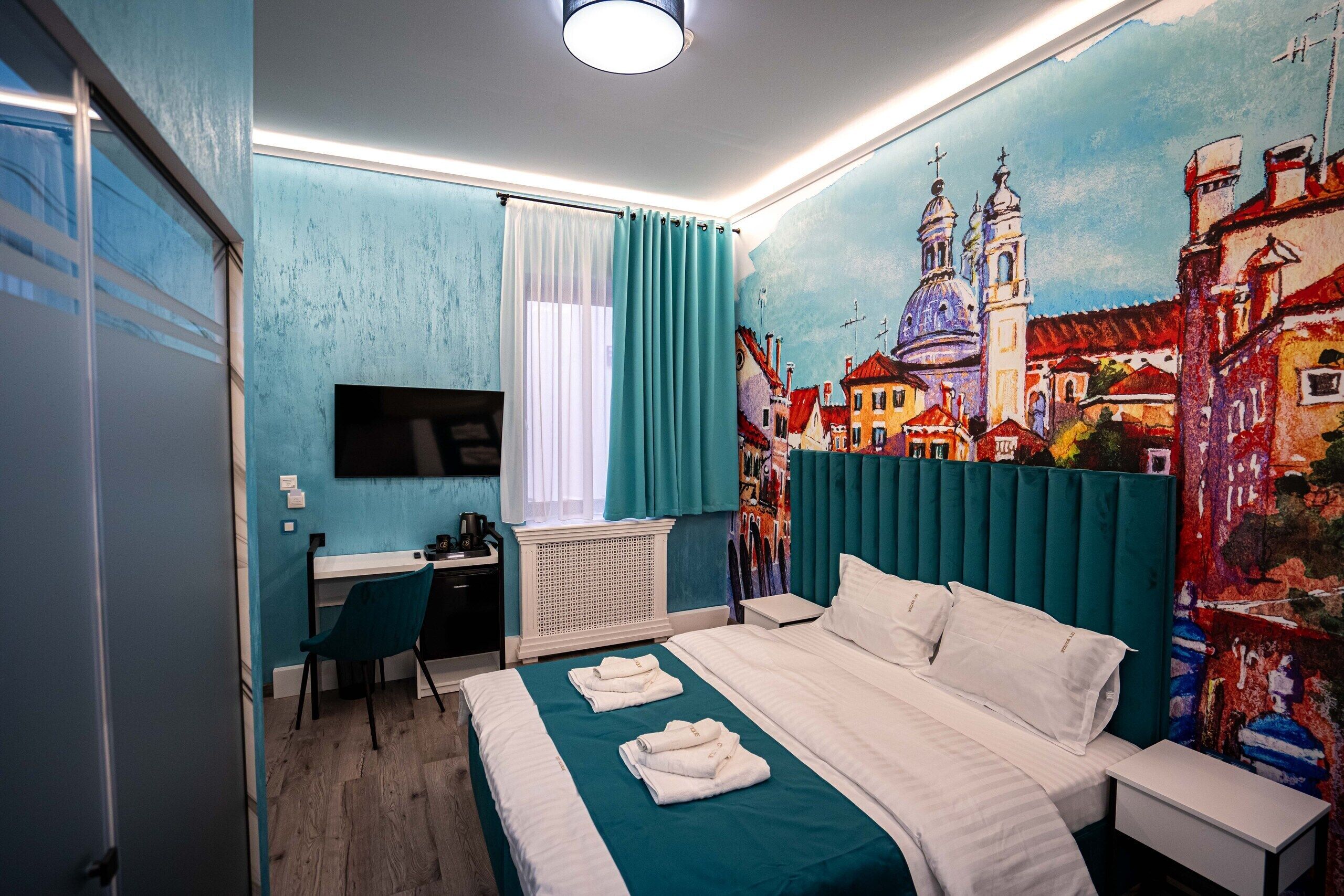 A hotel room with a bed, a desk, a chair, a TV, and a mural of a cityscape on the wall.