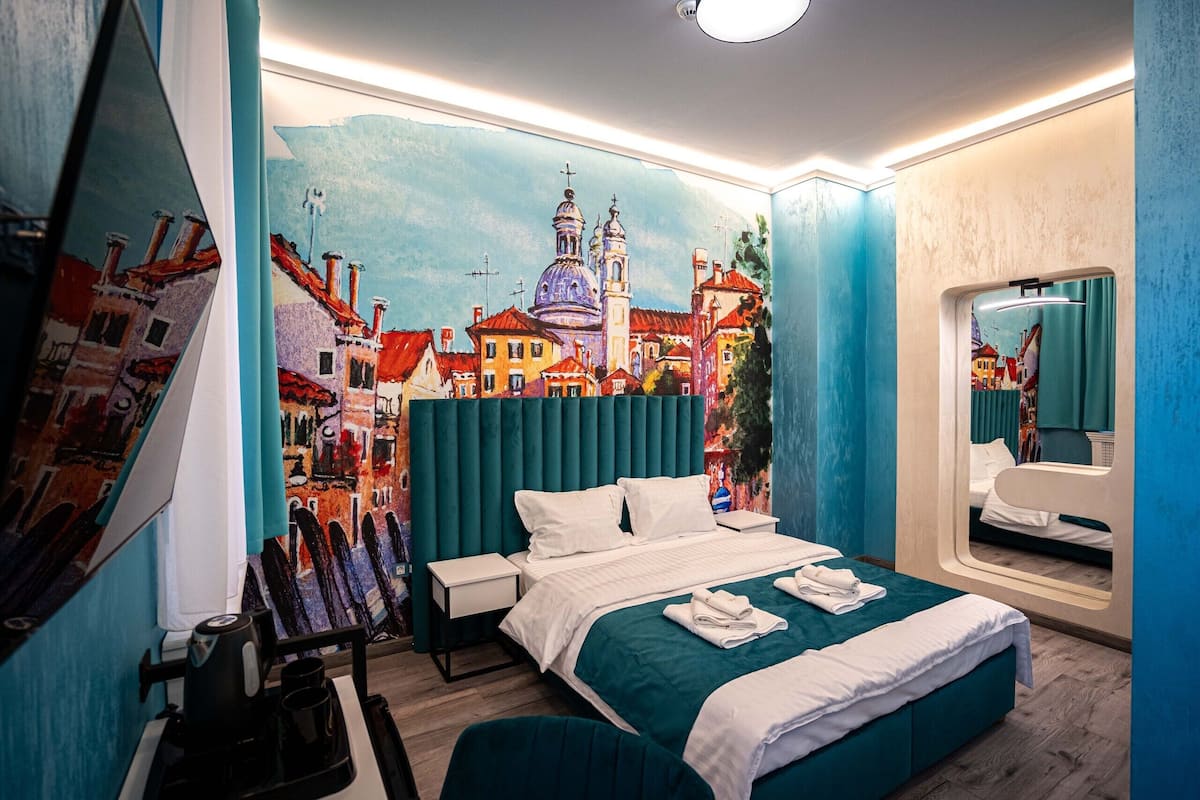 A modern hotel room with a large bed, a desk, and a painting of a cityscape on the wall.