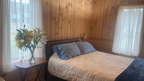 Een kamer met houten lambrisering, een bed, een klein tafeltje met een bloevas en een raam met dunne gordijnen.