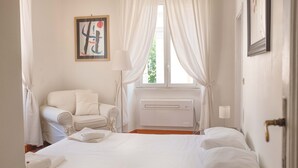 Een hotelkamer met een bed, een stoel, een raam met gordijnen, een radiator en een schilderij aan de muur.