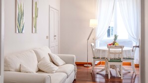 Een woonkamer met een witte bank, een eettafel met een stoel, een vloerlamp en een raam met gordijnen.
