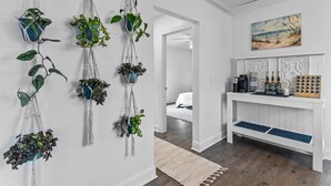 A hallway with macramé wall hangings, a painting of a beach, and a white console table with bottles and a coffee maker.