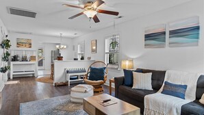 A spacious living room with a dark brown sofa, a white armchair, and a wooden coffee table. A ceiling fan and a chandelier are visible.