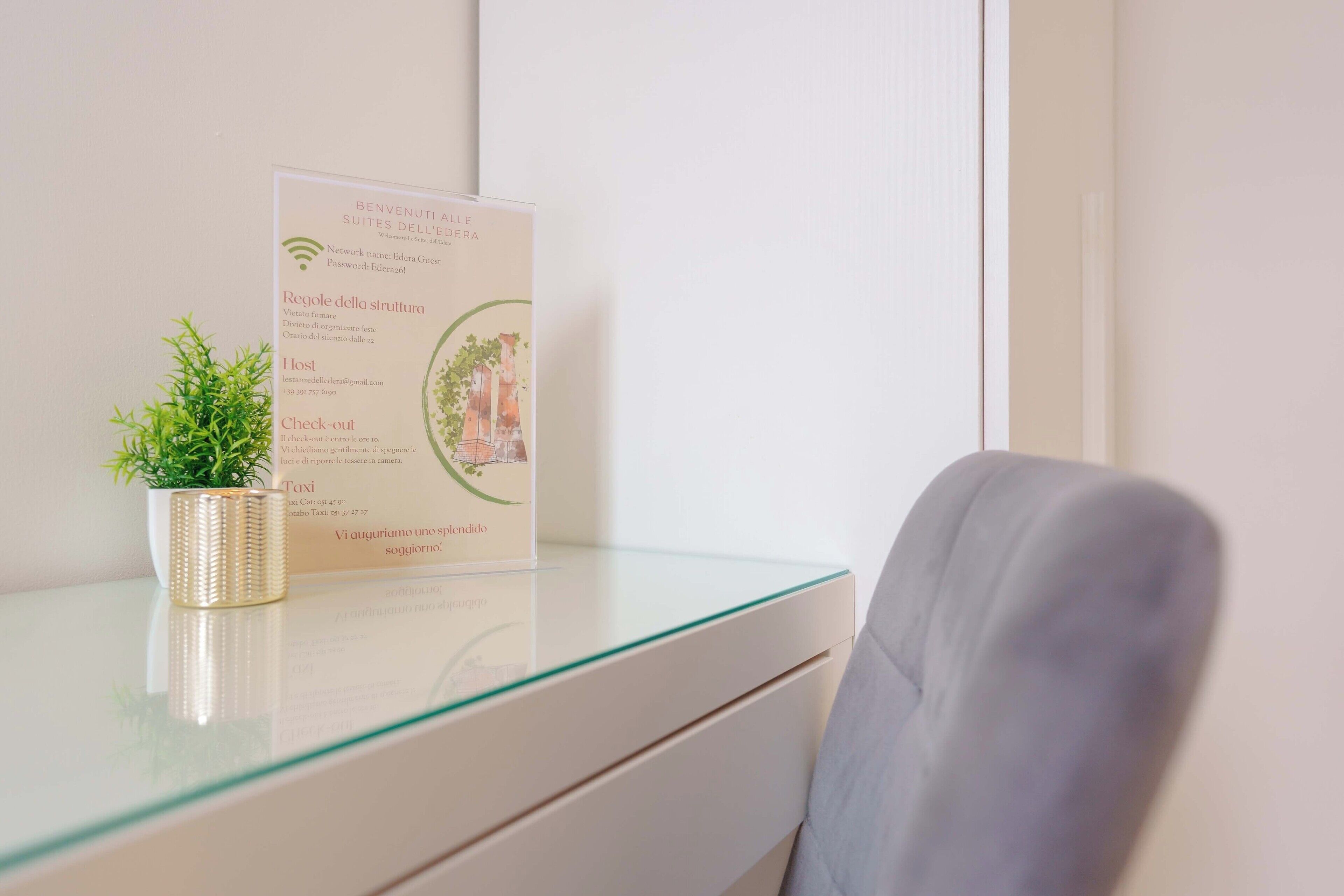 A hotel reception desk with a glass top, a potted plant, and a welcome sign detailing room rates and amenities.
