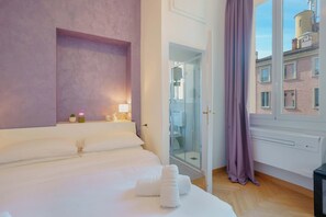 Ένα δωμάτιο ξενοδοχείου με ένα κρεβάτι, ένα μπάνιο με καμπίνα ντους από γυαλί και θέα στα κτίρια που βρίσκονται έξω.