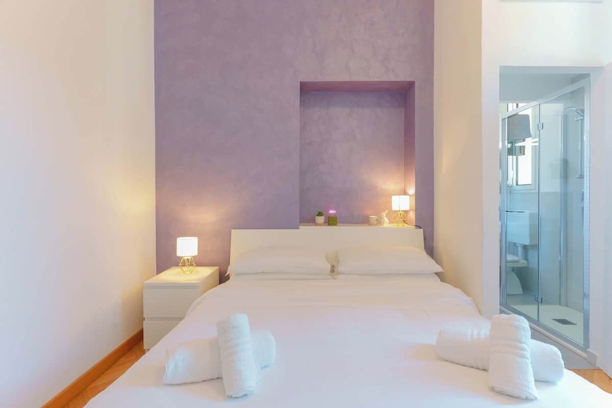 A hotel room with a large bed, two bedside lamps, a glass shower enclosure, and a purple accent wall.