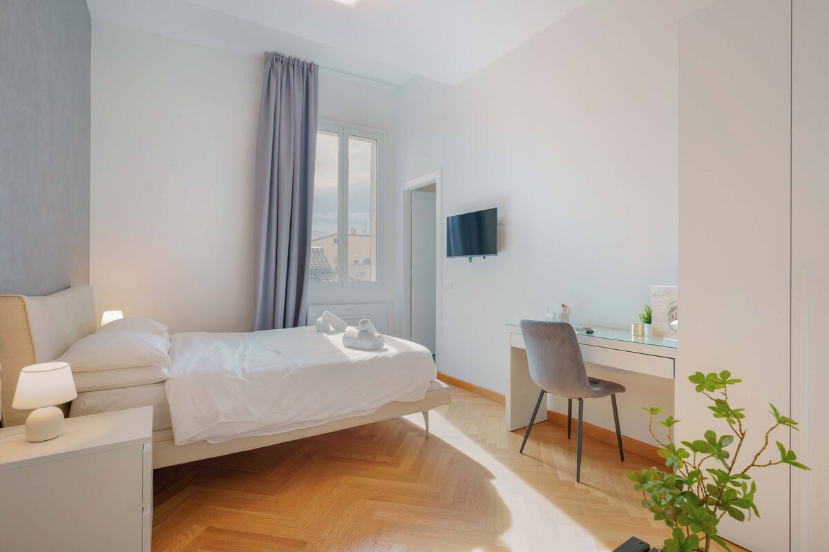 A modern hotel room with a bed, bedside table, desk, chair, and a TV on the wall.