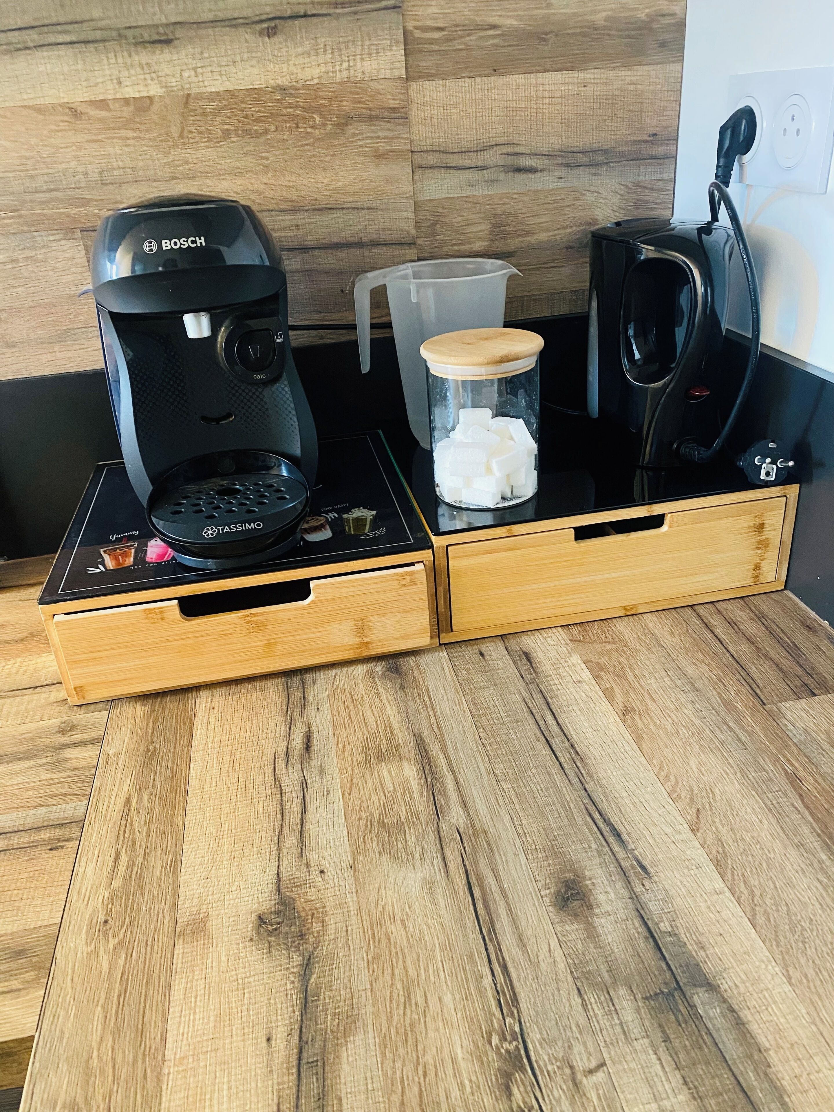 A kitchen counter with a Bosch coffee maker, a Tassimo pod holder, a glass jar with sugar cubes, and a black electric kettle.