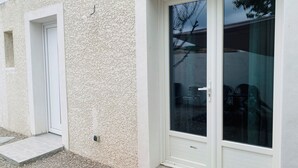 Fachada de edificio con pared blanca texturizada, una puerta blanca y un ventanal corredero que muestra muebles de exterior.