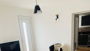 Sala de estar con televisor sobre un mueble de madera, una mesa de centro, dos sofás grises, una silla negra y un área de cocina visible a través de una puerta abierta.
